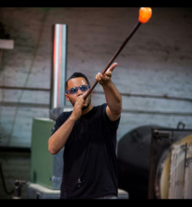 Leo Tecosky lifting and blowing a piece of hot glass. Symbiotic Spheres exhibiting artist.