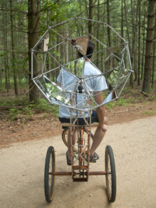 Nate Ricciuto riding a three-wheeled bike with an added umbrella of mirrored triangles, creating a camouflage with the trees.