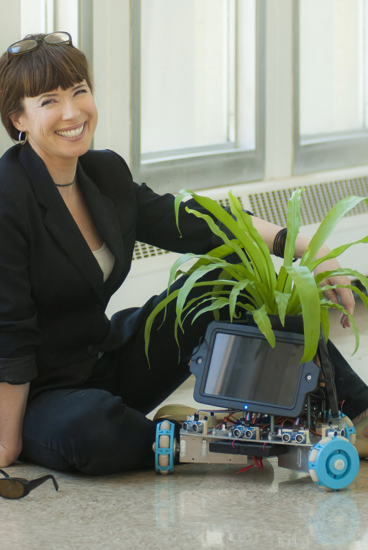 Elizabeth Demaray smiling, sitting with a small plant on top of a wheeled mechanism with a tablet screen, Symbiotic Spheres exhibiting artist