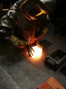 Amy Rubinstein in a full protective suit holding two hands on a red hot piece of glass.