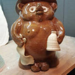 Ceramic bear with a wide brim hat, holding a bottle in one hand and a napkin in the other. Created by Tessa Carlton.