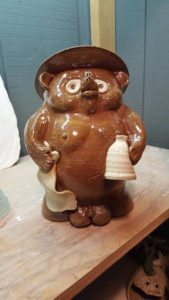 Ceramic bear with a wide brim hat, holding a bottle in one hand and a napkin in the other. Created by Tessa Carlton.