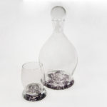 Clear glass decanter and a small glass, both with a slight purple tint with a flower motif. By Skitch Manion
