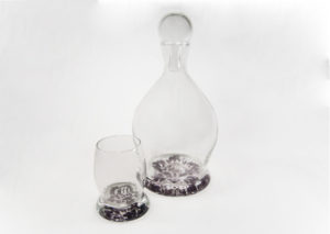 Clear glass decanter and a small glass, both with a slight purple tint with a flower motif. By Skitch Manion