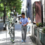 Video still of a man covered in small hexagonal mirrors walking next to a man on a city sidewalk. Created by Stanislav Müller.