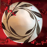 A white bowl, with large black leaves and stems winding around the top of the bowl, sits in front of a crimson background and branches of red berries. Created by Dorrie Papademetriou