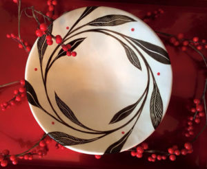 A white bowl, with large black leaves and stems winding around the top of the bowl, sits in front of a crimson background and branches of red berries. Created by Dorrie Papademetriou