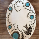 An oval platter with a black bird and blue flowers with black stems winding throughout the platter. Created by Dorrie Papademetriou