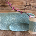 Purple flowers sit in a white ceramic cup with a soft blue band with wavy patterns carved in, sitting in front of two matching blue platters. Created by Dorrie Papademetriou