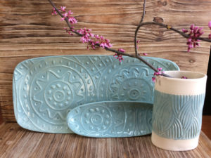 Purple flowers sit in a white ceramic cup with a soft blue band with wavy patterns carved in, sitting in front of two matching blue platters. Created by Dorrie Papademetriou