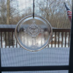 A glass medallion with an ornate cross design by James Sharpless, hanging on a screen window in front of a snowy yard.