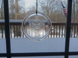 A glass medallion with an ornate cross design by James Sharpless, hanging on a screen window in front of a snowy yard.