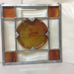 A glass panel with a crossed design and amber colored stained glass in each corner and a large amber color circle in the middle by James Sharpless