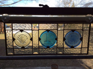 A glass panel with three circular glass medallions inside depicting a lighthouse, a shell, and a sand dollar by James Sharpless