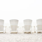 Photograph taken by Barry Hollritt of four white Adirondack chairs on a light tan beach.