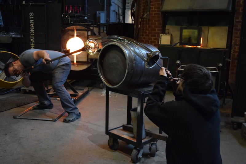 WheatonArts Glass Artists Skitch Manion and Brian R. bend and shape a large glass tube over a black barrel.