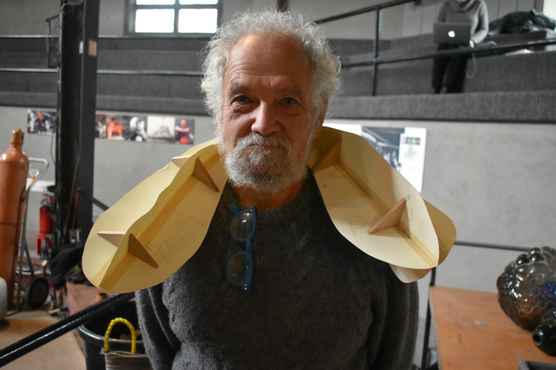 "Emanation 2019" Artist Allan Wexler smiling in a grey sweater with a yellow paper structure draped around his neck.