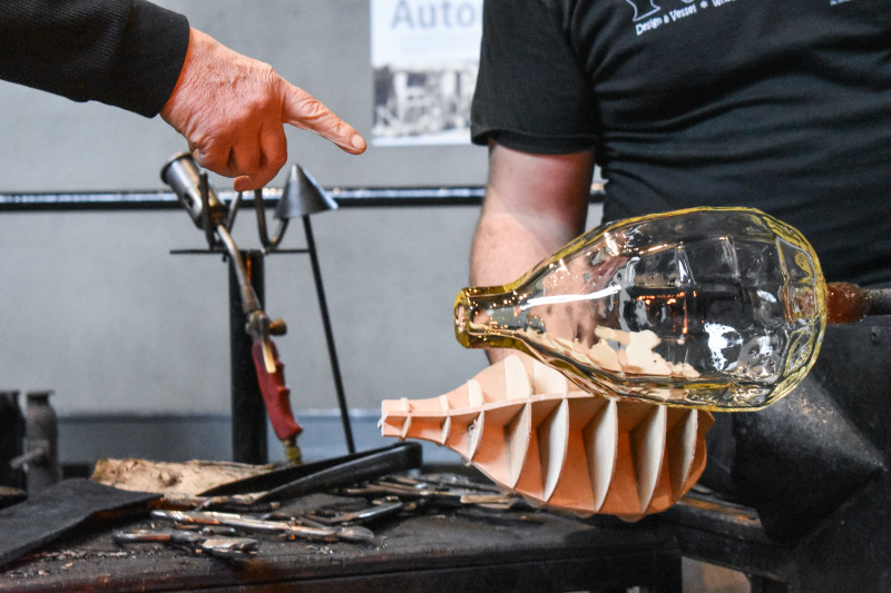 Closeup of "Emanation 2019" Artist Allan Wexler pointing at a newly created glass bottle at the end of a blowpipe.