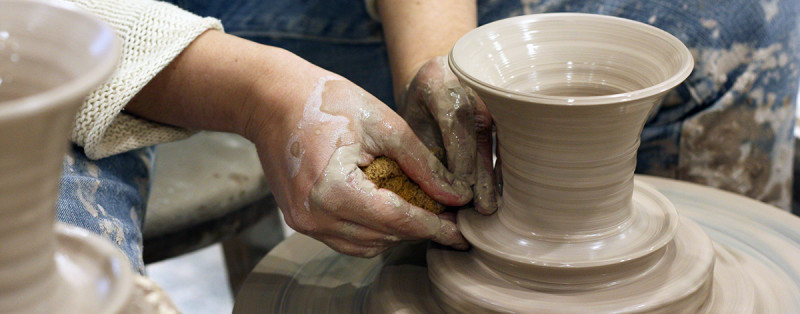 Woman spinning pottery wheel, using clay covered fingers to form the base of a vase