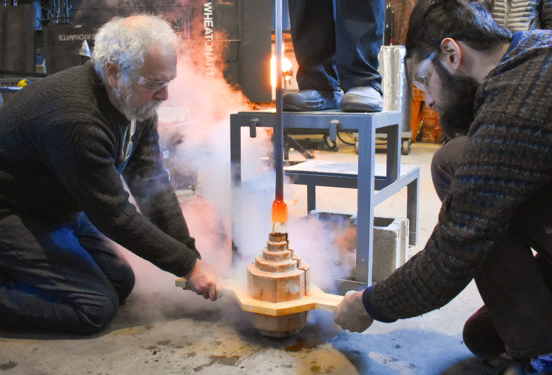 Emanation 2019 Artist Allan Wexler and WheatonArts Assistant Glass Artist Max Hertzan hold a mold in place while molten glass is poured in