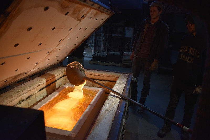 Closeup of a metal ladle pouring molten glass into a plaster mold of a rectangular door panel.