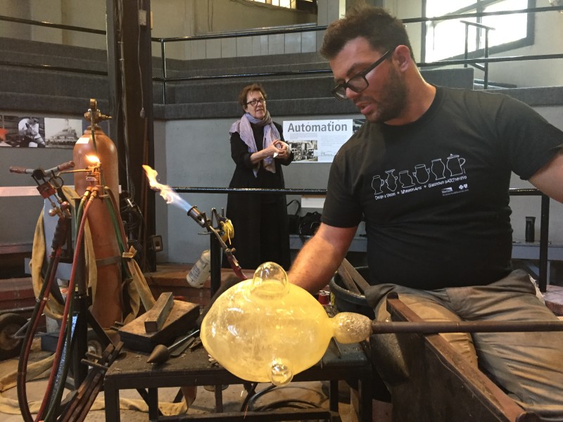 "Emanation 2019" Artist Jo Yarrington interacts with WheatonArts Glass Artist Skitch Manion as he creates a large, yellow glass orb for "The Uranium Game".