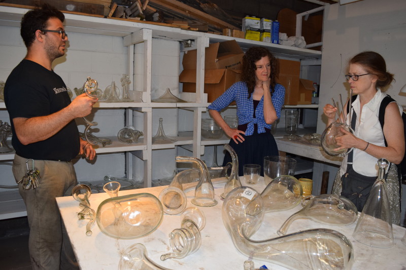 "Emanation 2019" Artists Laura Baird and Martha McDonald talk to WheatonArts Glass Artist Skitch Manion among a large collection of glass funnels, tubing, horns, and containers.