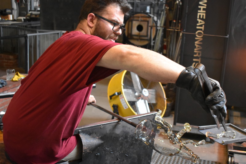 WheatonArts Glass Artist Skitch Manion fuses clear glass limbs to a clear glass bottle body.