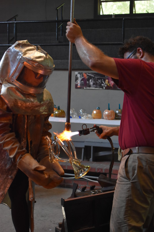 A Glass Artist in protective gear assists Skitch Manion as he uses a hot torch to shape a glass horn.