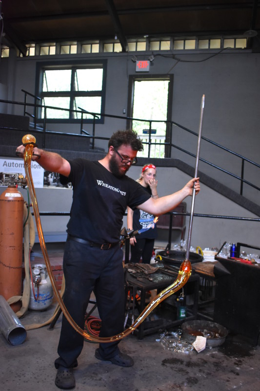 WheatonArts Glass Artist Skitch Manion pulls a long strand of hot glass, stretching from arm to arm.