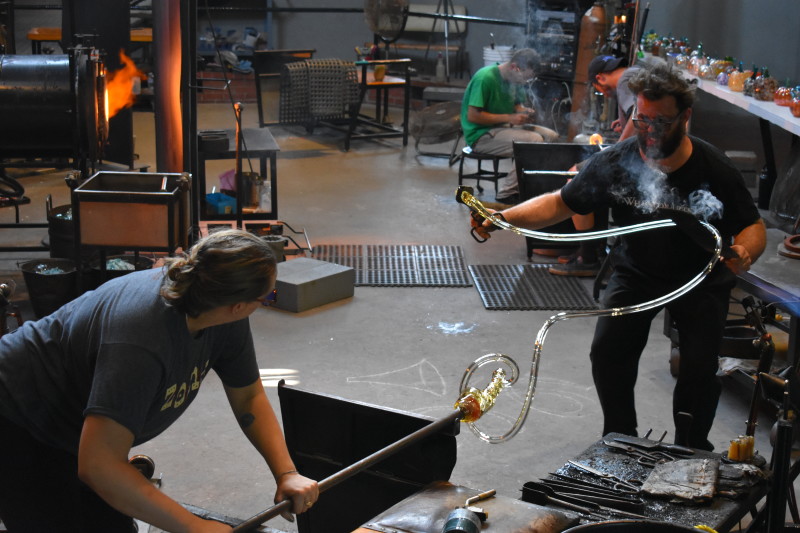 Two WheatonArts Glass Artists stretch and shape a long tube of clear glass.