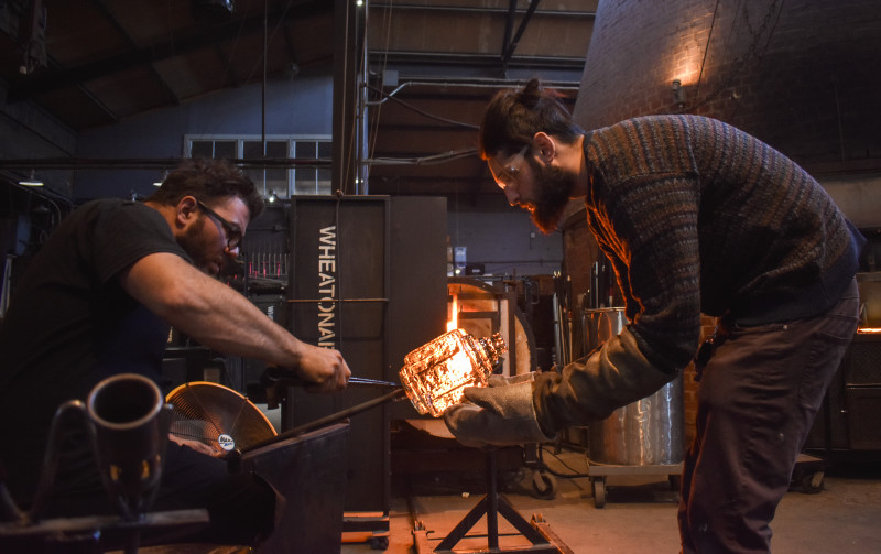 WheatonArts Glass Artist Skitch Manion and Assistant Glass Artist Max Hertzan carefully refining a red hot glass piece released from its mold.