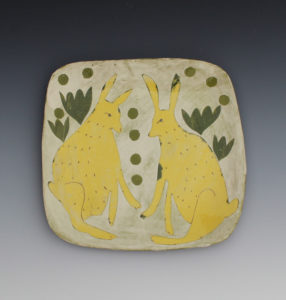 A white ceramic slab depicting two yellow rabbits with green leaves behind them. Created by Priscilla Dahl.