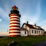 A square of a sharp photograph of a lighthouse with red and white stripes and a house behind it. The sky is blue with orange and yellow clouds in the far distance. Photographed by James Evangelista
