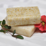 Two offwhite bars of soap displayed on a soft white sheet with a single red flower running around the bars. Created by Bay Berry Bliss