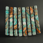 Glass piece by Stephanie Baness made up of 8 columns colored in a marbled pattern of orange, teal, and white, connected with 8 rows of black behind.