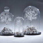 Three flameworked clear glass trees of various sizes by Stephen Brucker with exposed roots, each contained under a glass cloche