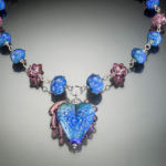 Piece of jewelry by Sharon Carlucci with a blue heart pendant and blue and purple alternating beads going up either side
