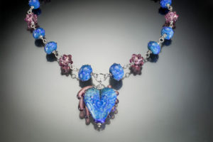Piece of jewelry by Sharon Carlucci with a blue heart pendant and blue and purple alternating beads going up either side