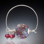 Piece of jewelry by Sharon Carlucci with a large multicolor circle as the focal piece and three dangling red glass beads.