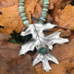Silver metal clay leaf pendant with turquoise beads of various shades, laid on a bed of orange autumn leaves. Created by Sue Fontana.