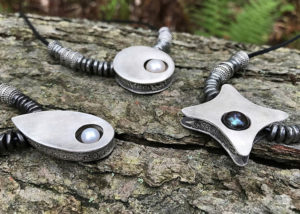 A closeup of three pendants in a teardrop, circle, and star shape, each with a pearl within, draped over a piece of wood. Created by Sue Fontana.