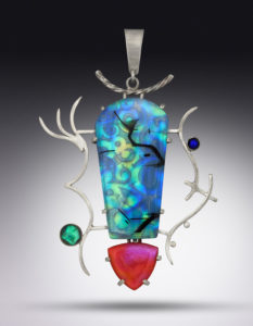 A silver pendant by Paul Lorber with a smooth blue and green gemstone decorated with swirling patterns, two small gemstones in blue and green at each side, and a deep red reuleaux triangle cradled at the base.