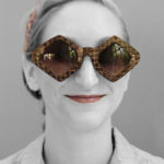 Black and white headshot of a person with a color pop on the brown diamond sunglasses by Laurie Olefson as well as the mahogany color lipstick and tan headband.