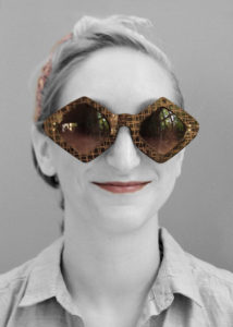 Black and white headshot of a person with a color pop on the brown diamond sunglasses by Laurie Olefson as well as the mahogany color lipstick and tan headband.