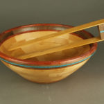 A pair of wooden tongs rest on a smooth wooden salad bowl with a deep brown trim and a thin blue ring. Both are created by John Baun.