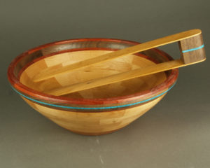 A pair of wooden tongs rest on a smooth wooden salad bowl with a deep brown trim and a thin blue ring. Both are created by John Baun.