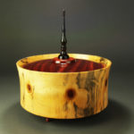 A wooden circle container with a deep red lid and black post on top. Created by John Baun.