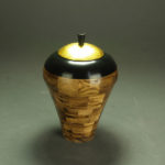 A vessel by John Baun made up of a wooden square pattern, a thick black rim, and a bright golden lid.