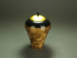 A vessel by John Baun made up of a wooden square pattern, a thick black rim, and a bright golden lid.
